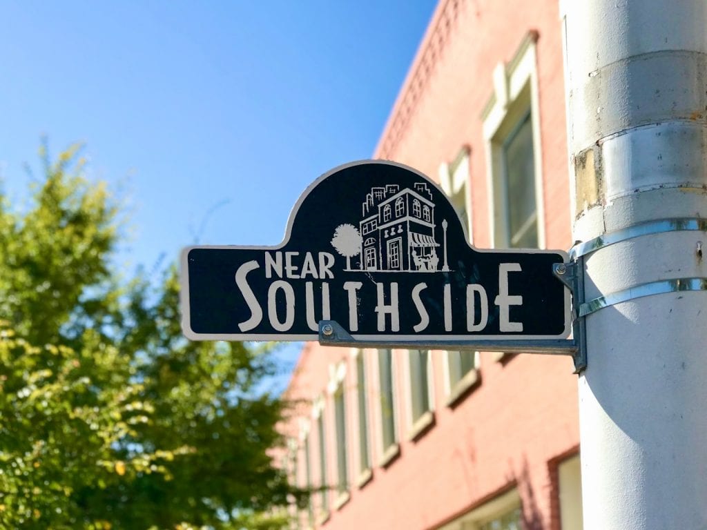Black street sign reads "Near southside" with an outline of a building, the sign is against a blue sky with a red building in the background