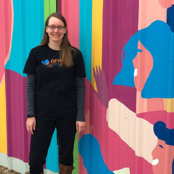 a woman wears a DFW Craft Shows shirt and poses in front of a long wall full of art that reads Good Vibes Only in Fort Worth Texas