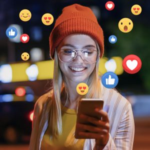 A woman in a beanie smiles at her phone while react icons float around her in the night sky