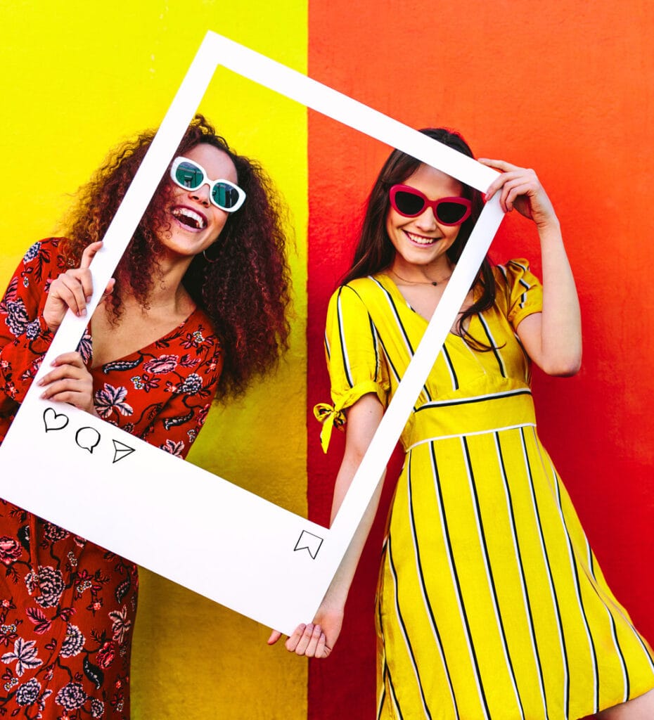 two women stand inside of a large photo frame prop with instagram buttons written on the bottom