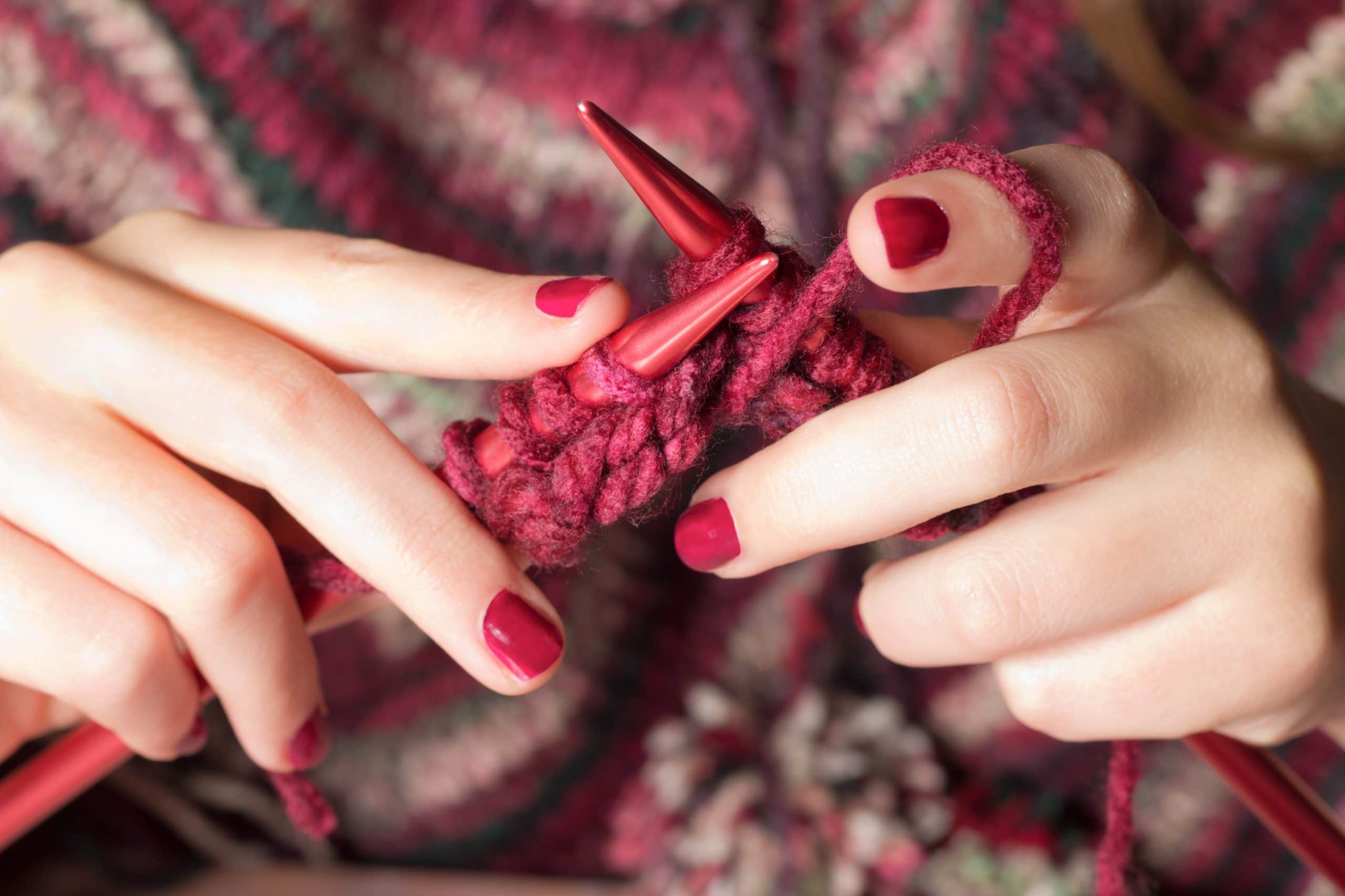 Two hands can be seen holding fuscia knitting needs that match their thread and finger nails. They are working on what looks to be the beginning of a project.