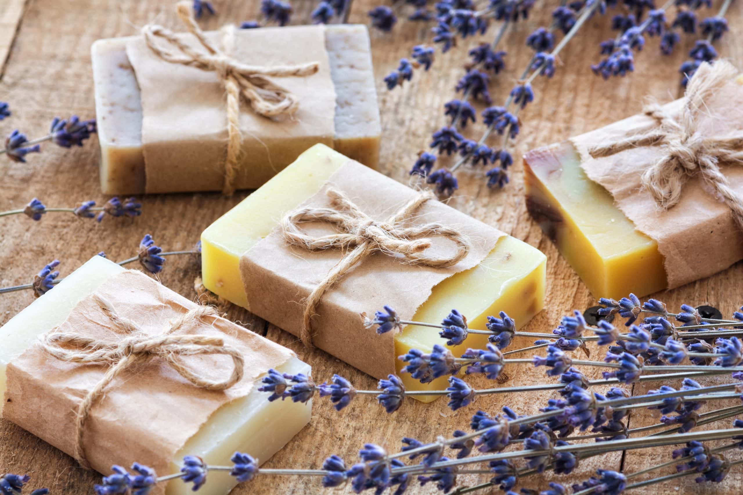 Several bars of handmade soap are packaged neartly with kraft paper around the middle and baker's twine wrapped into bows. There are lavendar springs all around the soap, which is sitting on a wood tabletop.