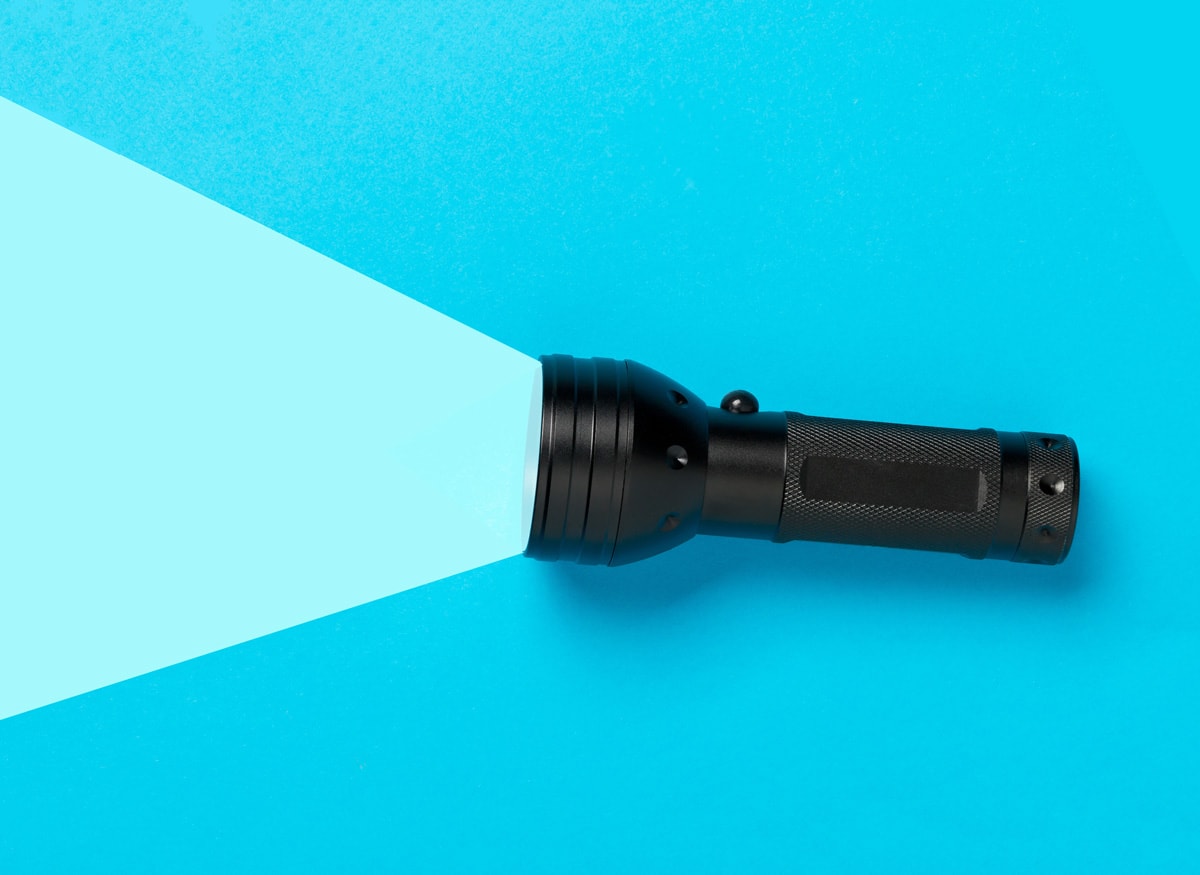 A flashlight is laying horizontal on the right side of the screen. The flashlight is black, and a light blue highlighted area is casting out of the flashlight as if it is lighting up the background. The other side of the background is a darker blue.