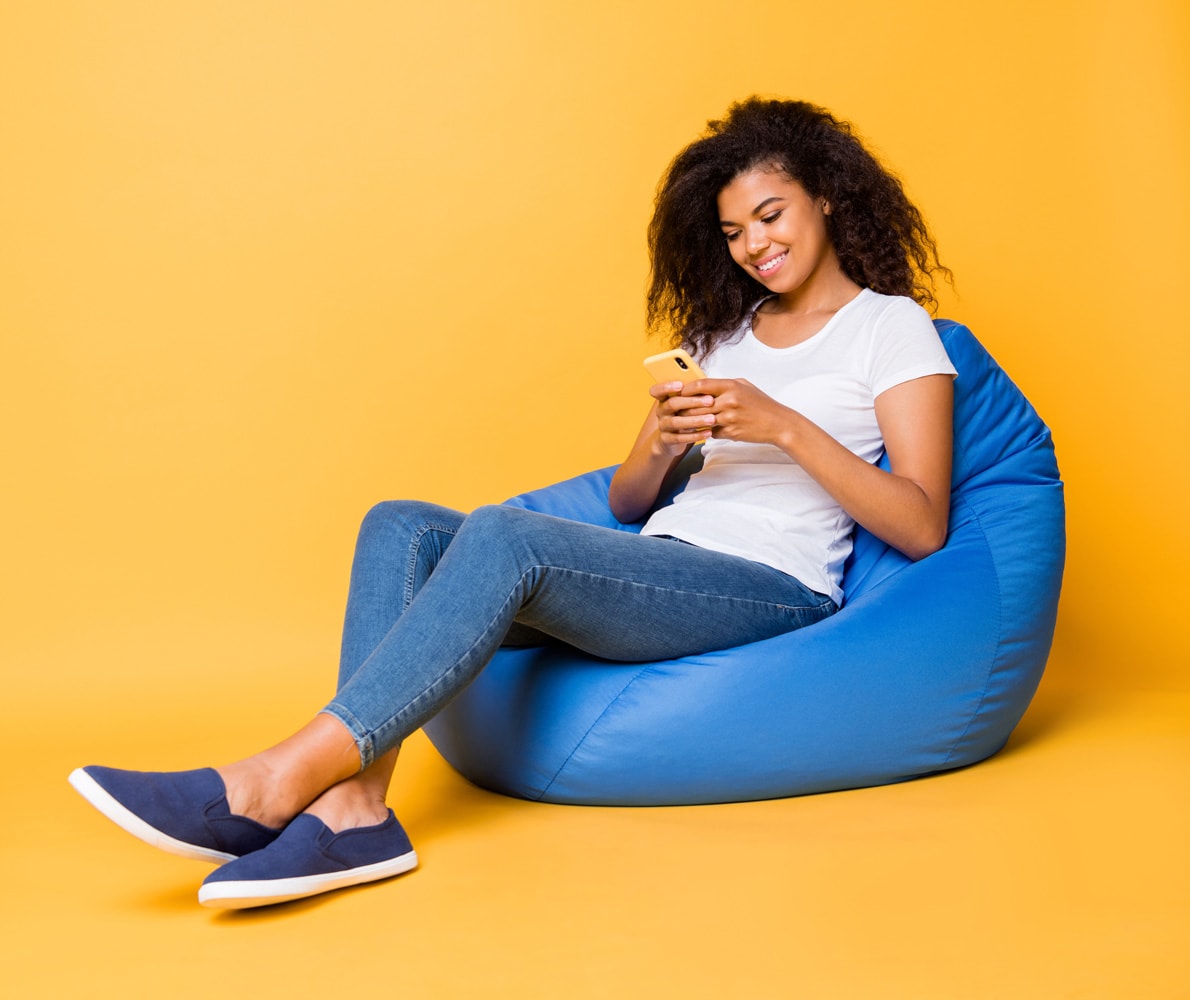 A woman of color sits on a bright blue bean bag wearing a white shirt and denim. She is looking at her phone. The background is a pale but still bright orange