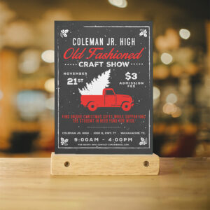 An event flyer sits in a blonde wooden perch on a blonde wooden table in front of light colored bokeh. The flyer is an eye catching design for the Coleman Jr High Old Fashioned Craft Show
