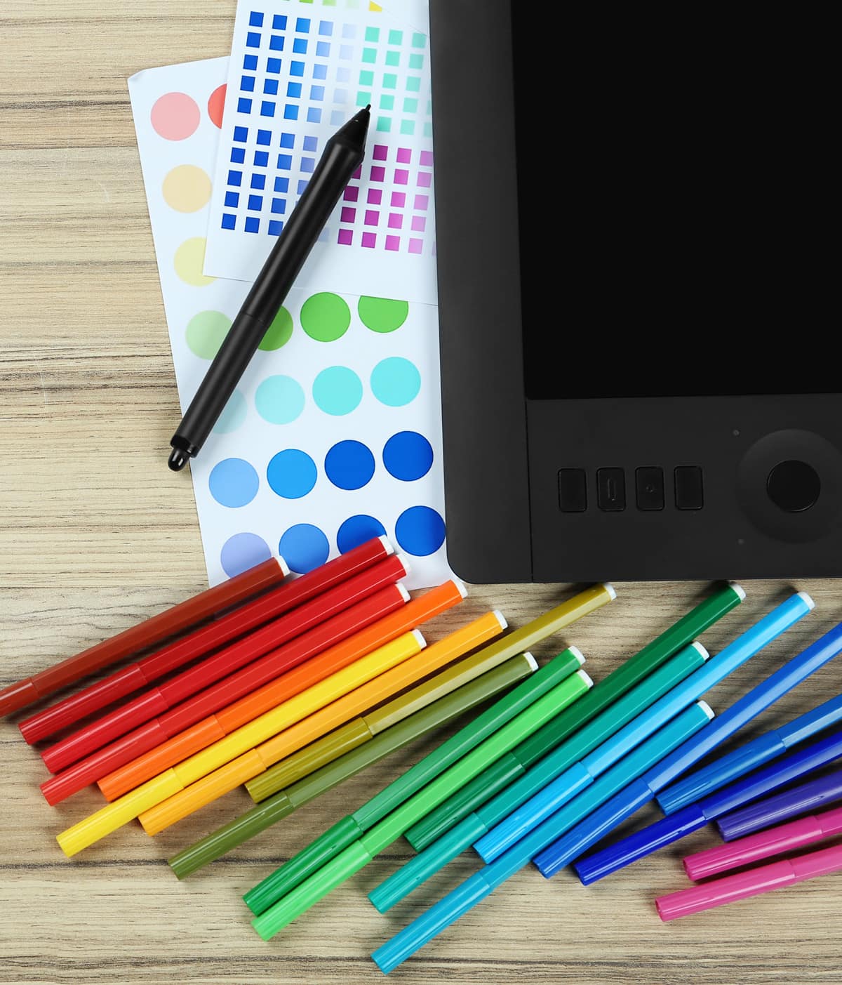 An array of colored markers lay on top of a sheet of color swatches. Everything is next to a design tablet with a stylus resting nearby.
