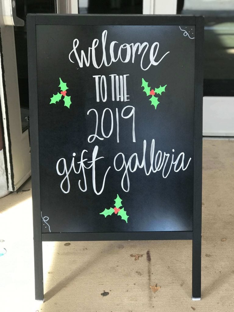 A chalkboard sign stands in front of an event with handwritten cursive text that says "Welcome to the 2019 gift galleria" with three mistletoe leaves and berries around it.