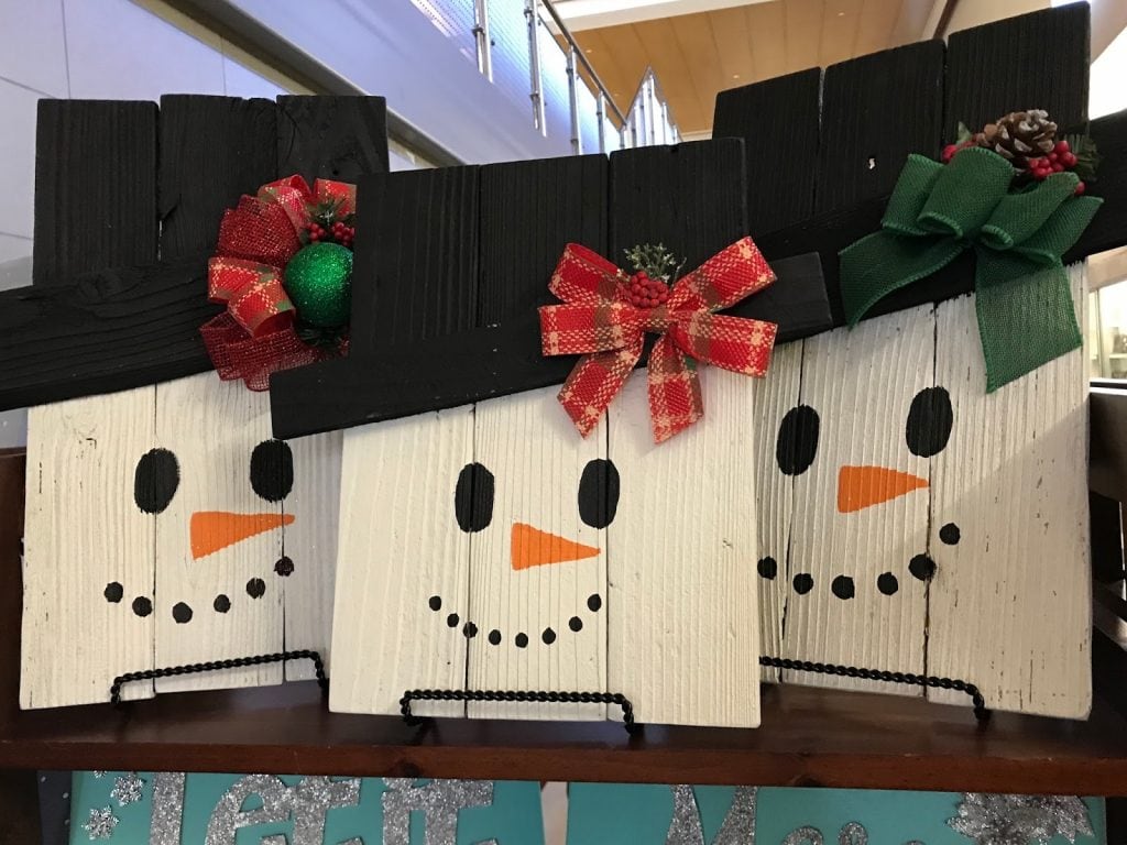 Three wooden panels joined together to create a rectangle on which a snowman's face has been painted. Each snowman has a bow or some kind of decoration on their hat.