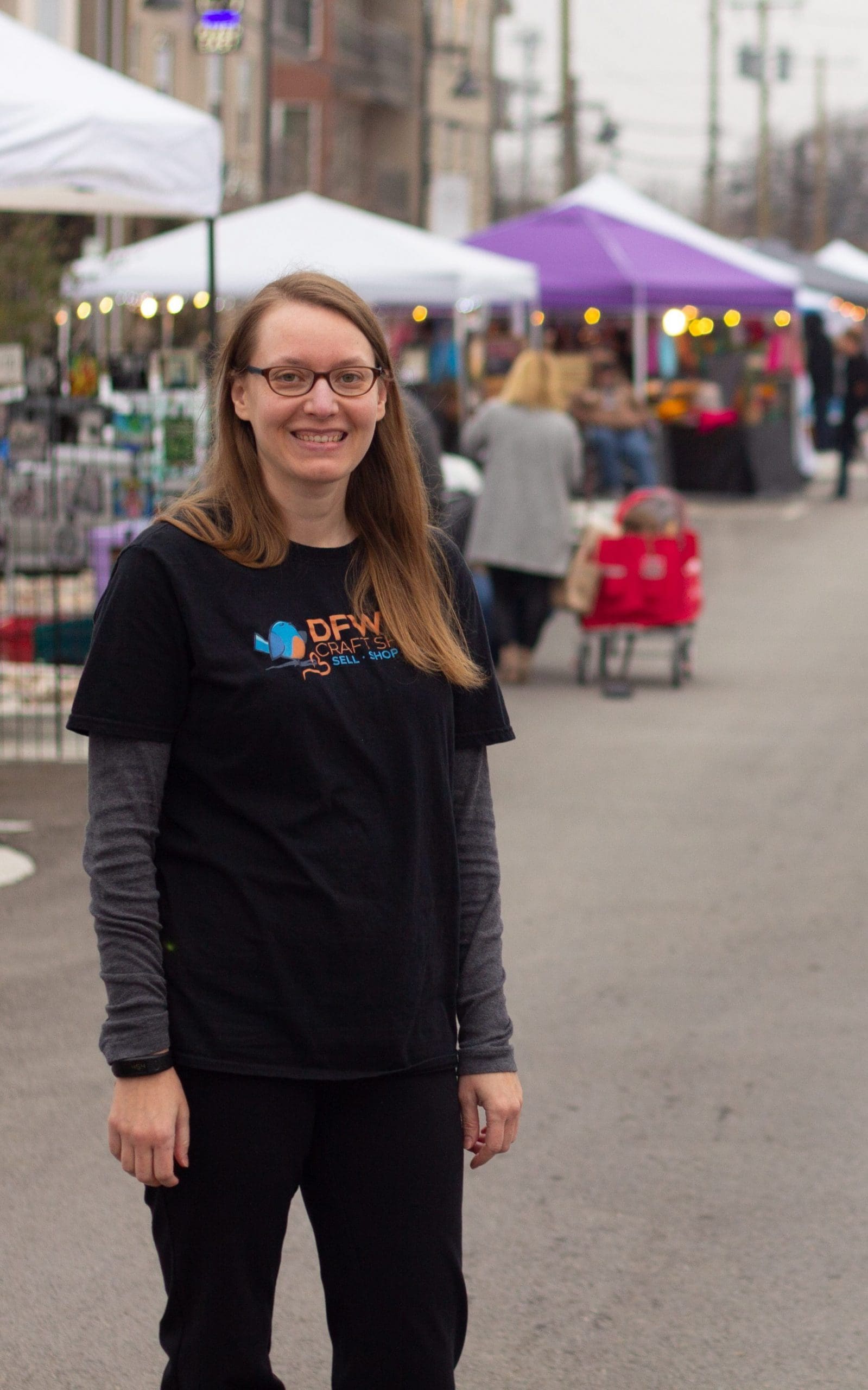 Tania of DFW Craft Shows stands at a street craft fair in Fort Worth Texas. She is wearing a DFWCS shirt and there are colorful canopies behind her.