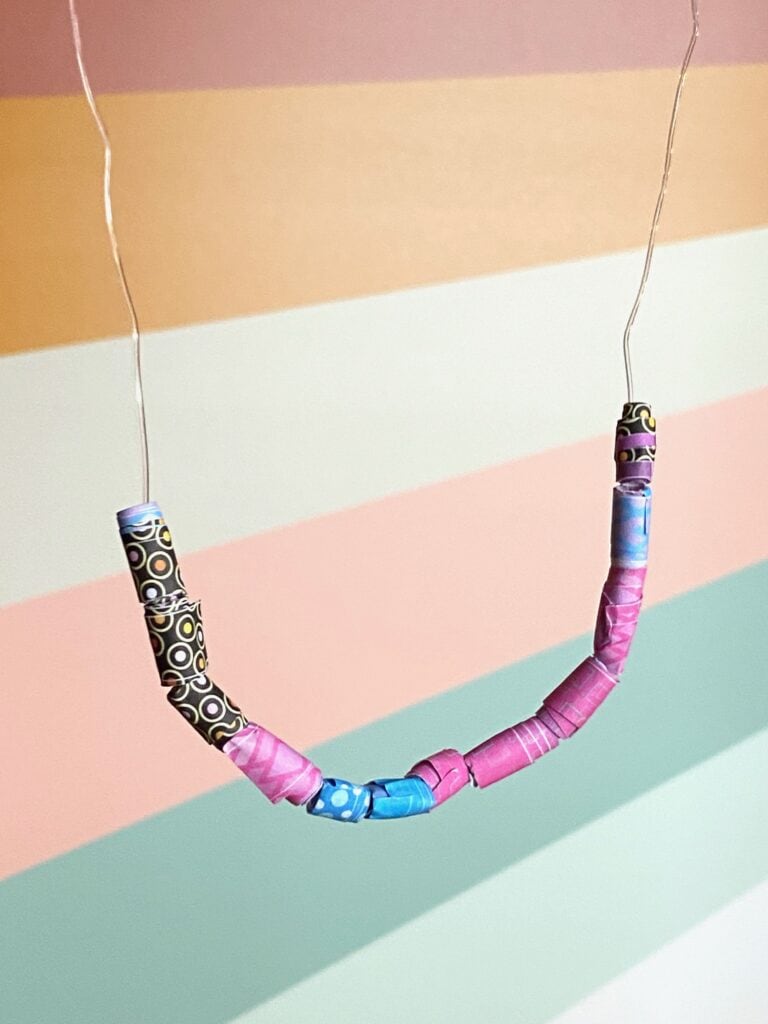 A finished necklace full of paper beads dangles in front of a colorful background.
