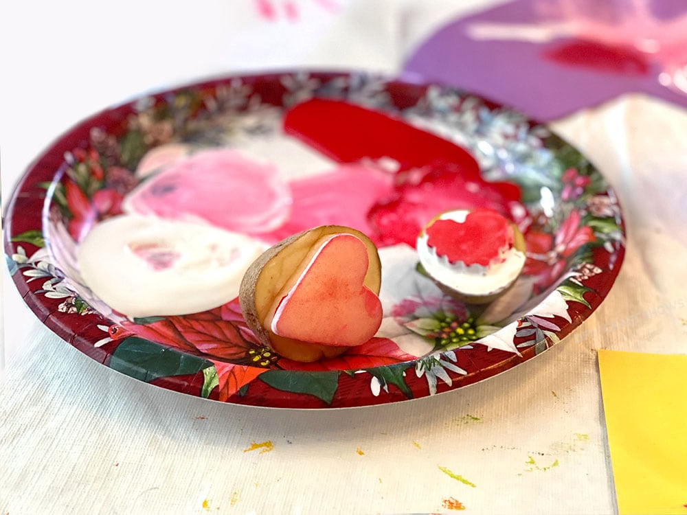 A potato carved into a heart stamp sits on a paper plate covered in red, white, and pink paint