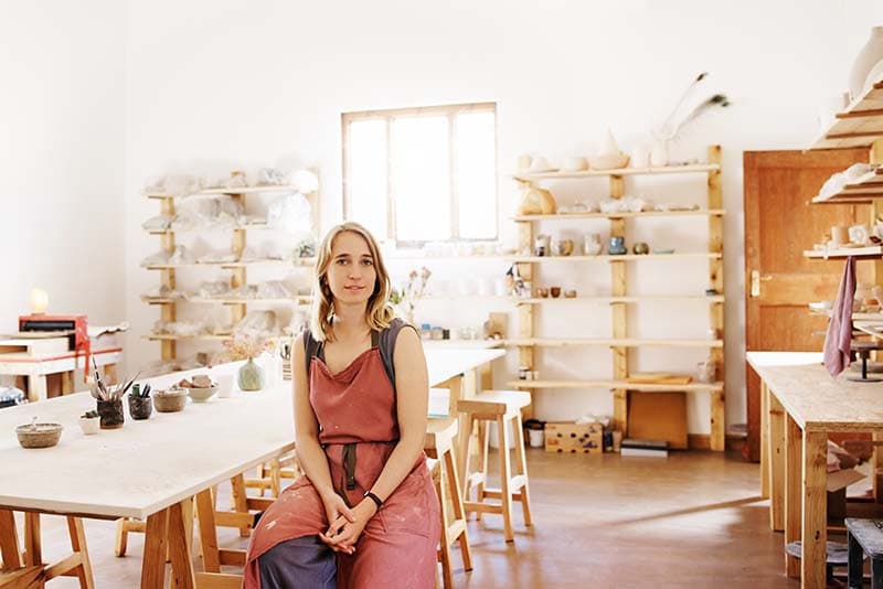 A woman in a pink apron sits at her empty work table. Shelves of finished art are behind her and vases line the table.  She has a slight, uncertain smile.