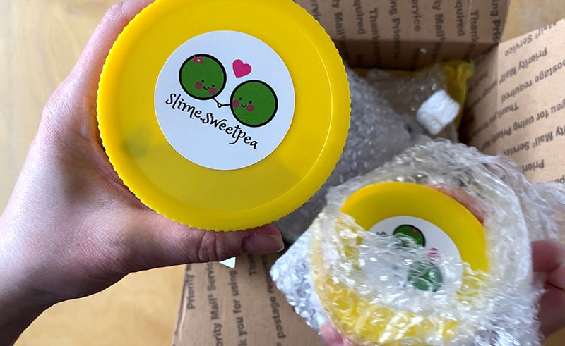 holding two slime sweetpea slime containers fresh out of the box. They are clear containers with yellow lids and a logo with two peas and a heart between them