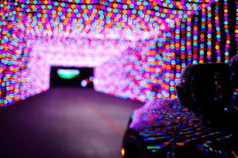 Purple, red, blue, and yellow lights shine through a long tunnel and reflect off the side of a car driving through