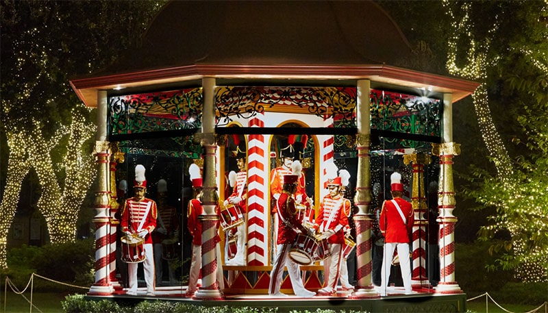 several people are dressed as drummers around a small gazebo at night.  The trees are covered in lights for the 12 days of christmas at dallas arboretum