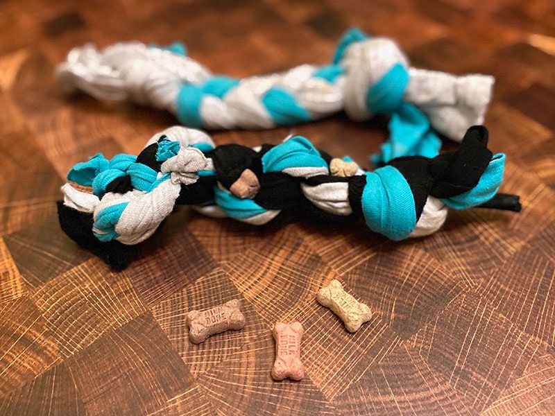 Two different dog enrichment toys lay on a wood surface, one with small mini milkbones tucked tightly into the weave of the braid or knots