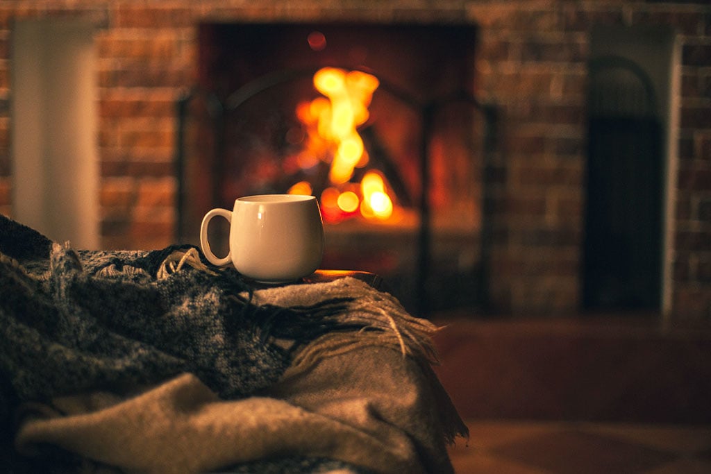A fire burns in the fireplace while a person cuddles up in blankets and a cup of hot cocoa