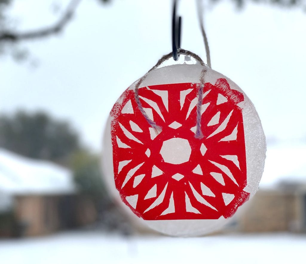  A red snowflake hangs in a round block of ice from a green tree. The background is covered in snow.