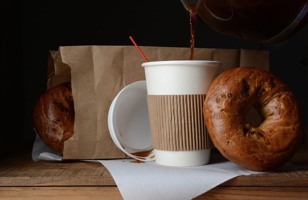 A cup of togo coffee sits on a napkin next to a large bagel with a paper bag in the background