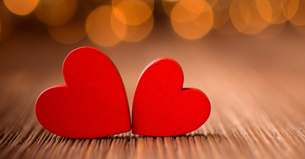 Two red wooden hearts sit on a wood grain table with bokeh lights in the background