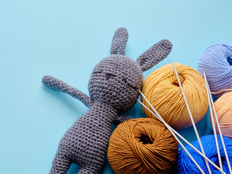 A gray knitted bunny sits next to a colorful array of cotton yarn balls in orange, yellow, and varying blues. Knitting needles lay across the balls of yarn. The background is a light blue.