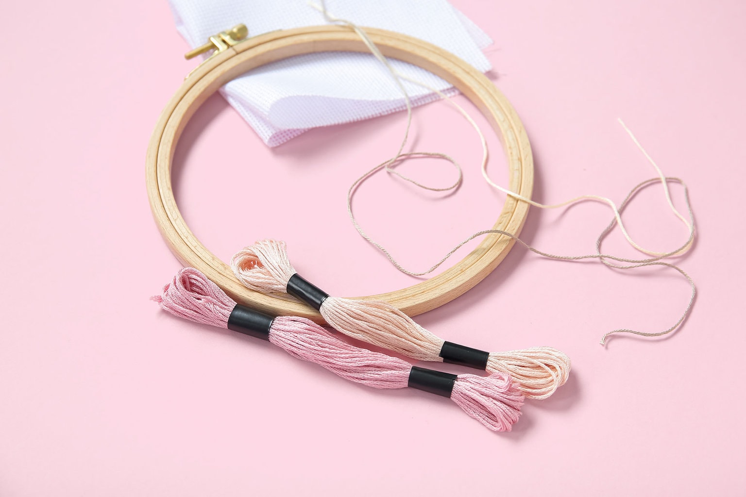 An empty embroidery hoop sits on a pastel pink background next to two shades of pink embroidery floss and white fabric