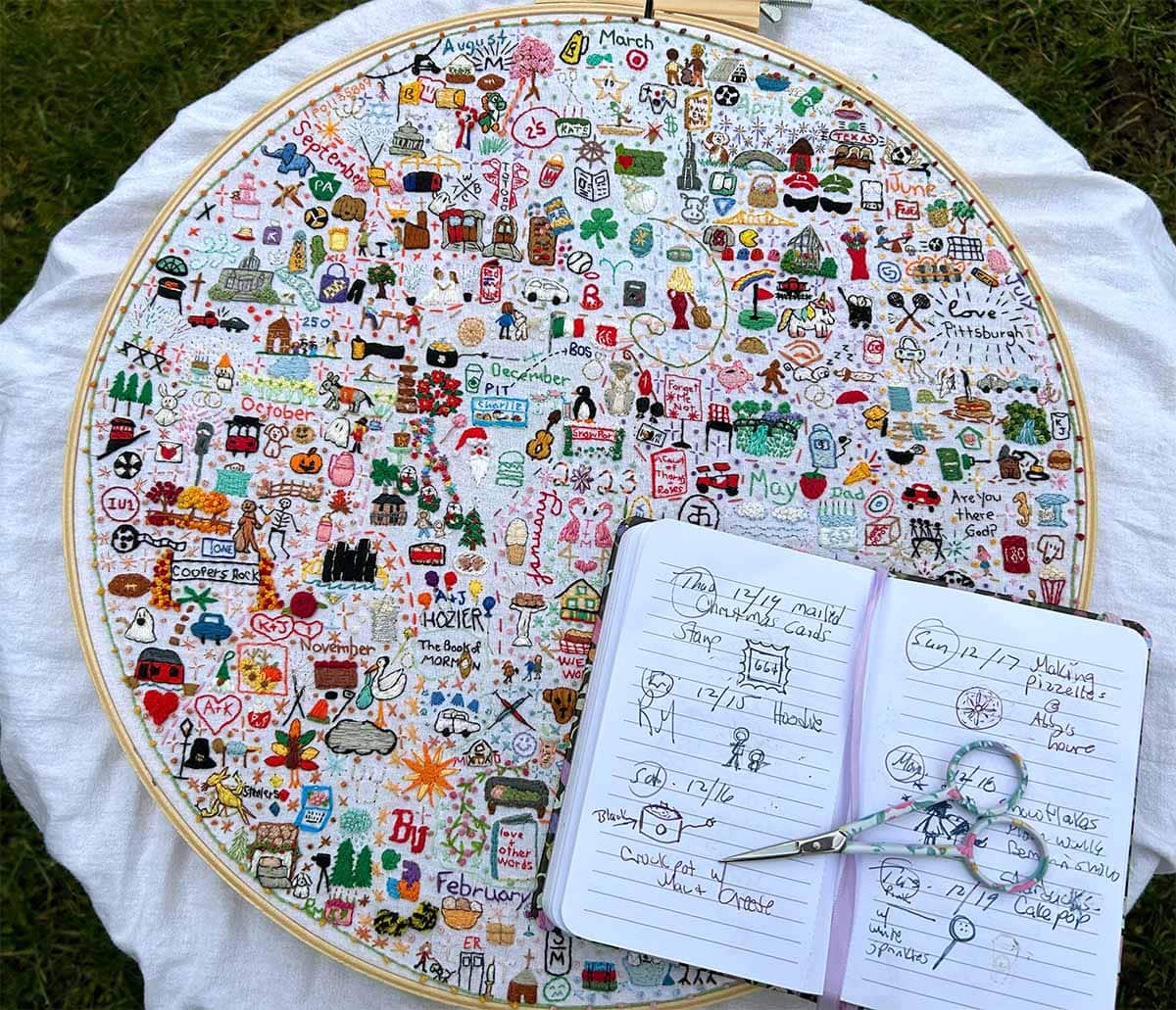 A 14"embroidery hoop is filled to the edge with embroidered icons. A design notebook is held open by embroidery scissors to a page with sketches for the hoop