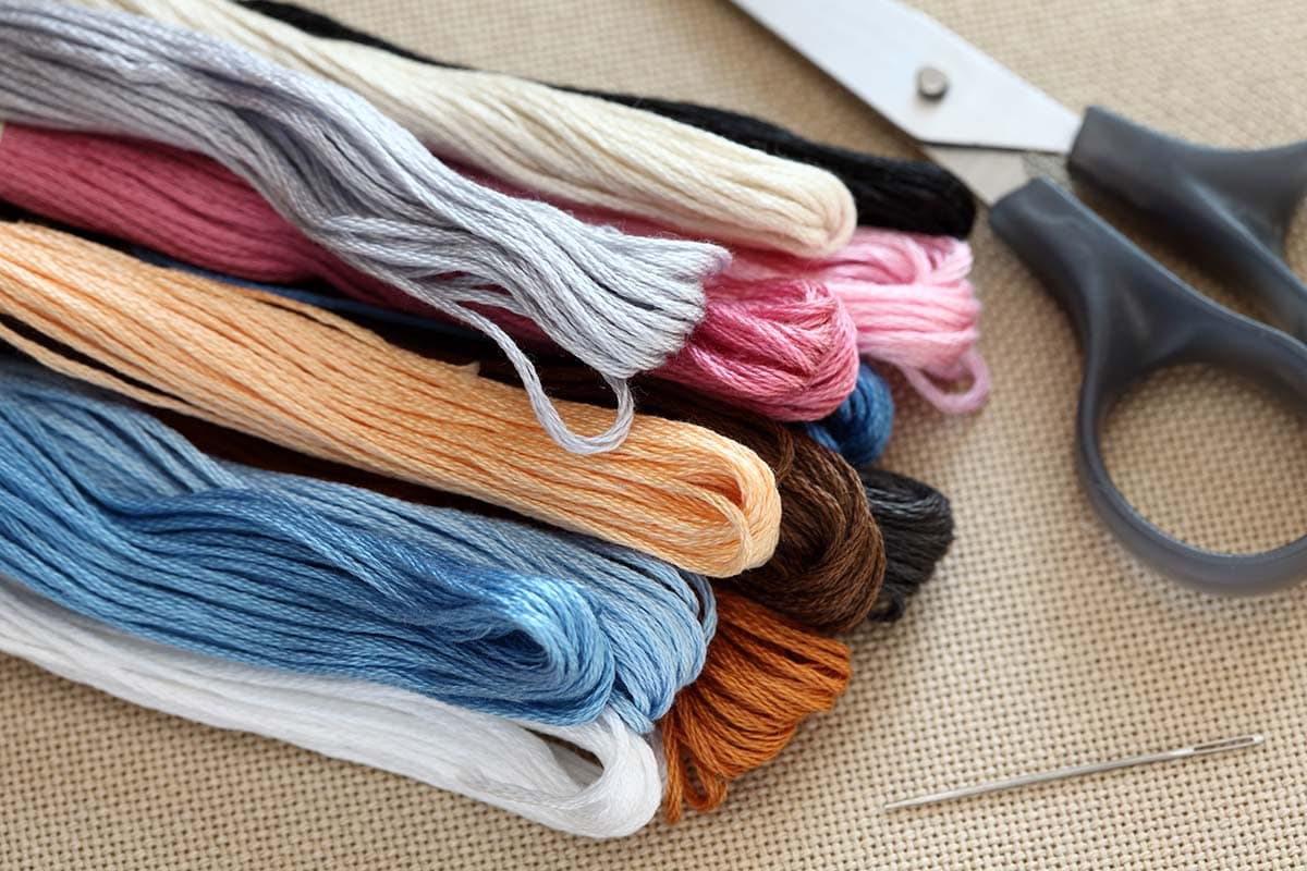 a stack of colorful embroidery floss sits next to gray handled scissors and an embroidery needle.  all piled on top of fabric