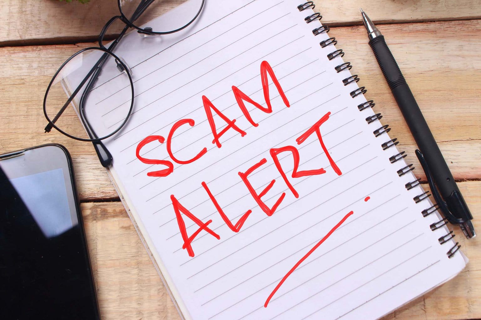 a white notebook lays beneath glasses and a pen. the notebook reads "scam alert" in red ink