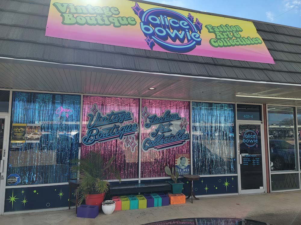 The sun glares off a sign that reads Alice Bowie with a yellow to pink gradient in the background.  The sign is on the front of a brick building with large front windows showing the inside of a thrift store.