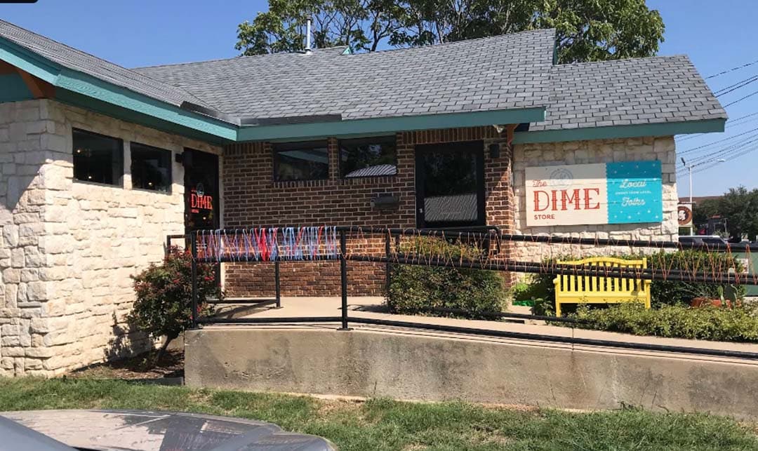 A building covered in white rock and red brick with a gray roof.  Wooden sign out fronts reads "The dime store."  A ramp entrance with yarn on the railing can be seen.