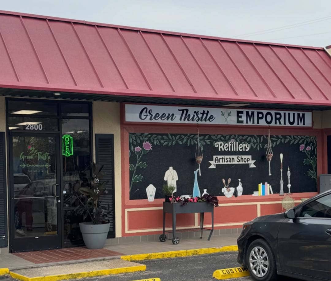 A red roofed building with a white sign that reads "green thistle emporium" with a cute boutique style mural on the front.