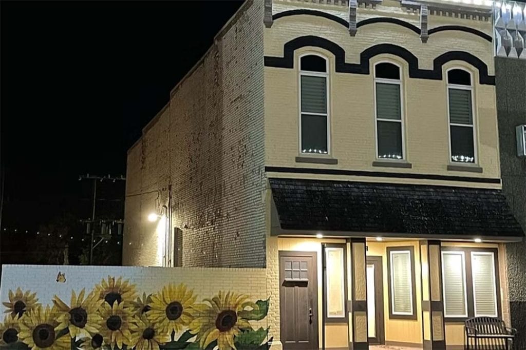 A two story building with yellow bricks is lit up against the dark night sky, the nearby brick fence is painted with sunflowers