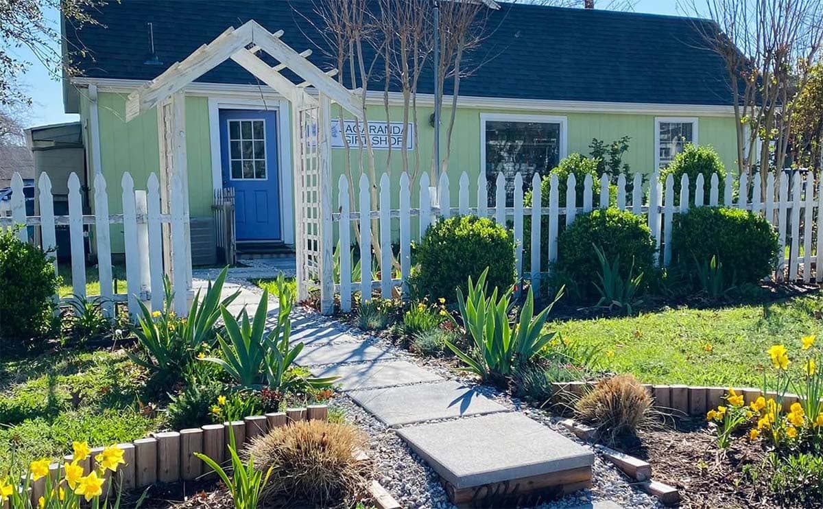 A beautiful garden stands before a pale yellow building with an archway over the sidewalk. Sign reads Jacaranda Gift Shop
