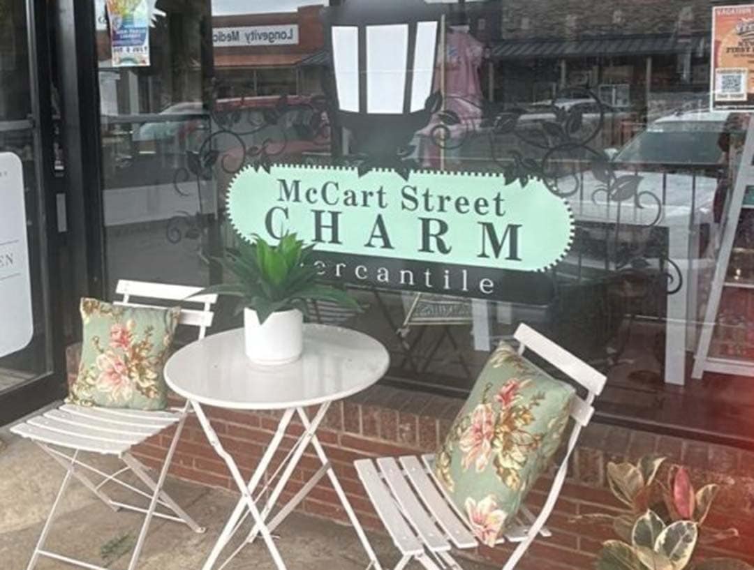 Two white patio chairs sit in front of a glass window with text that reads "McCart Street CHARM Mercantile"
