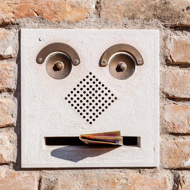 A metal mailbox is imbedded in a pale beige brick wall. The mailbox has hardware that looks like eyebrows, eyes, nose, and a mouth. There is a letter sticking halfway out of the mouth.