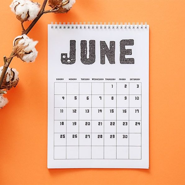 A white calendar is open to the "June" page against an orange background. There are white flowers laying diagonally across the left side of the screen.