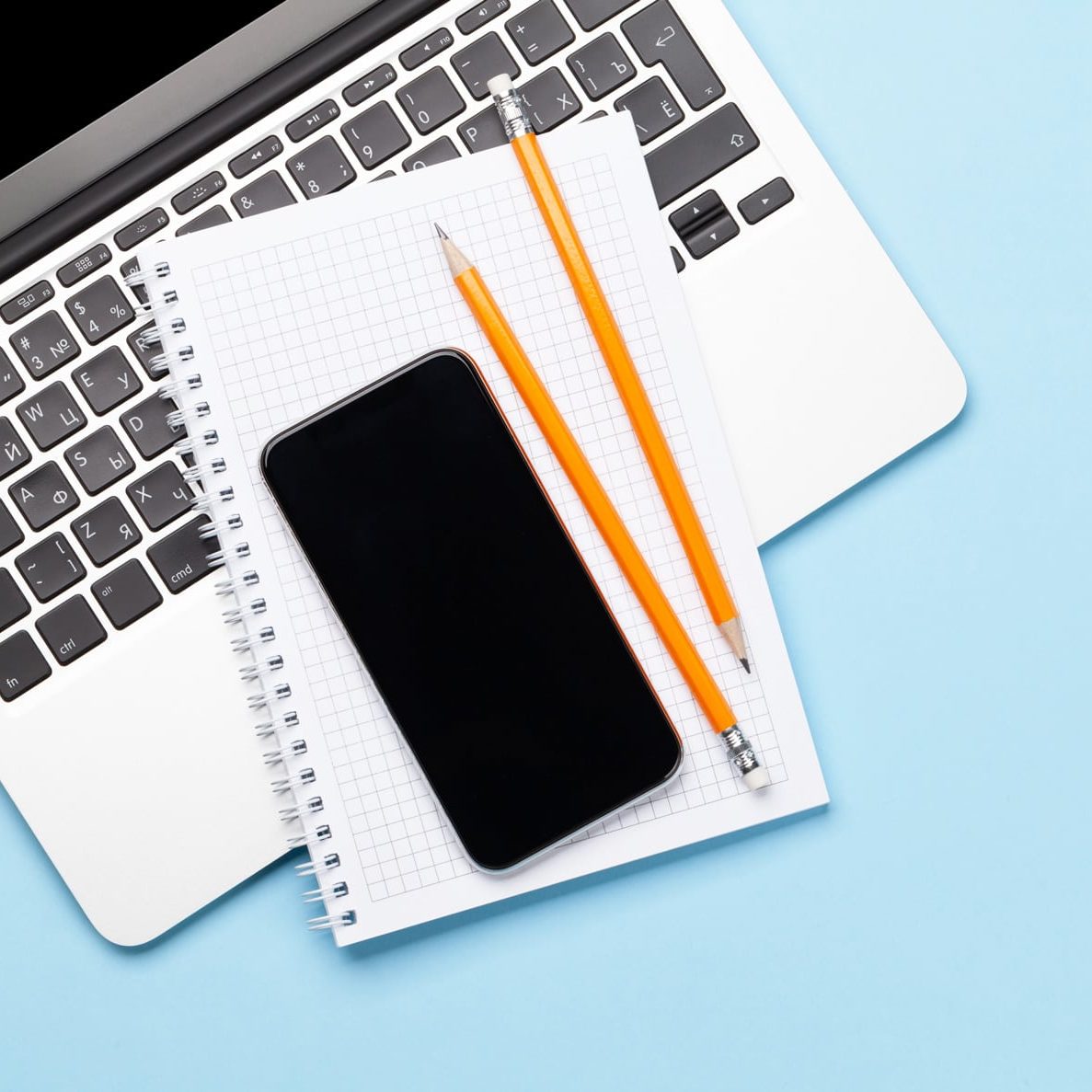 A spiral notebook with two orange pencils lays on top of an open laptop. There is a black cellphone on top of the notebook.