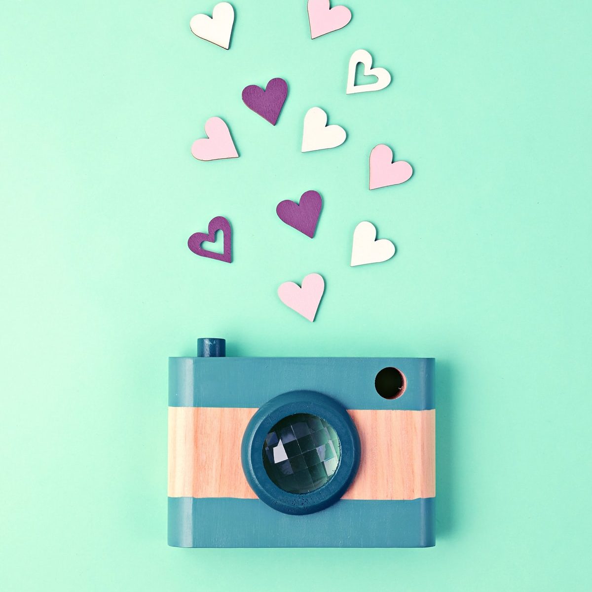 A cute, green and beige, wooden camera is posed with several pink and purple wooden hearts coming out of it. They are arranged on a mint background.
