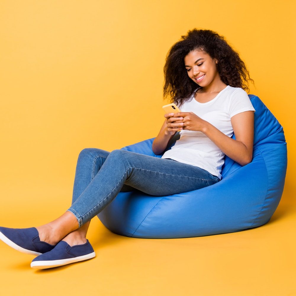 A woman of color sits on a bright blue bean bag wearing a white shirt and denim. She is looking at her phone. The background is a pale but still bright orange